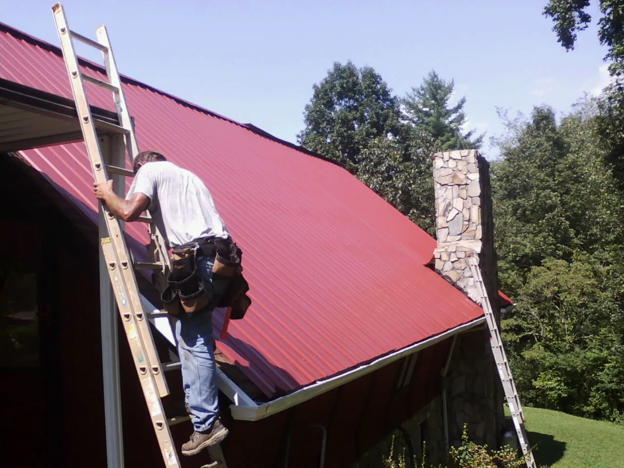 Roofer on a ladder beside a red metal roof and stone chimney — Soffit & Fascia Repair by Roofing Brothers in Stow